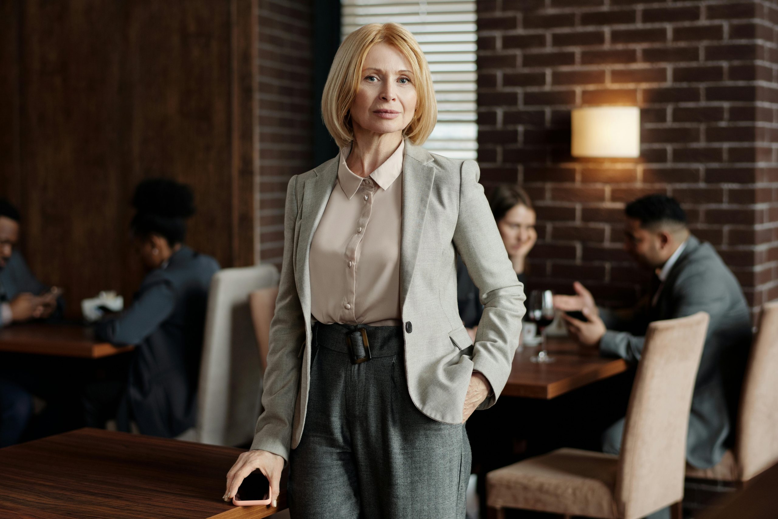 Confident businesswoman in formal attire standing in a modern café with colleagues working in the background.