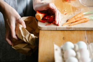 Unrecognizable female putting seeds of red bell pepper from cutting board into carton package while cooking at counter with carrots and eggs