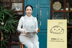 A woman holding a tablet sits outside a Japanese dessert cafe with a welcoming smile.