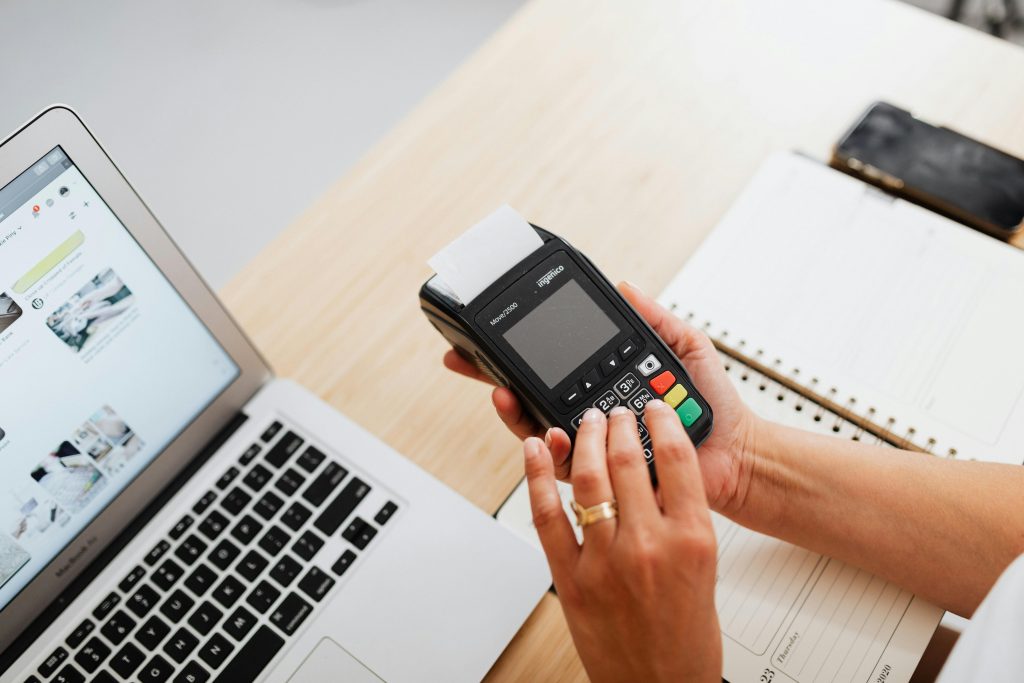 A person using a payment terminal with a laptop and a notebook on a work desk, illustrating cashless transactions.