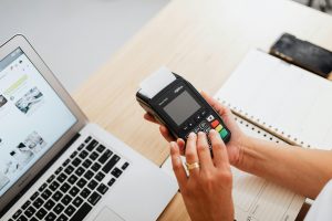 A person using a payment terminal with a laptop and a notebook on a work desk, illustrating cashless transactions.