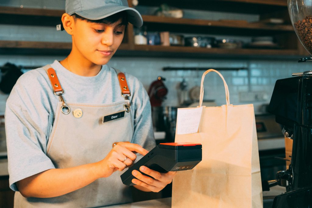 A young barista processes a transaction using a wireless terminal in a cozy cafe setting.